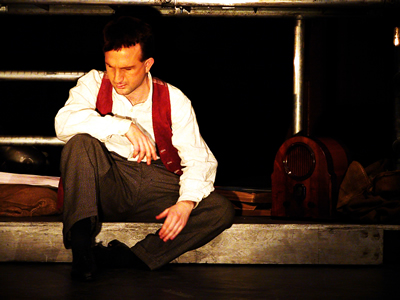 Younger Jacques, sitting on lower level of scaffold, with knees up, elbow resting on them, listening intently to 30s style backerlite radio