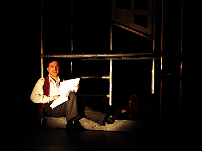 Younger Jacques, sitting back against lower level of scaffold pole, with one leg stretched out and one knee bent, holding up the brailed book he is reading.