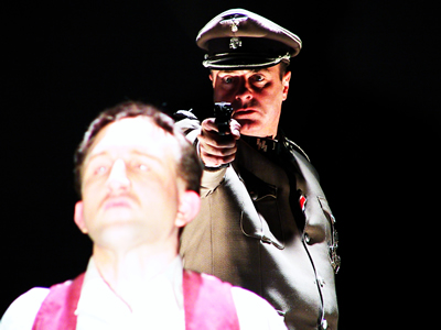 Major in background, from waist up, in Nazi grey military uniform, pointing a gun straight out at shoulder height towards Younger Jacques in foreground, brightly lit, facing out with head slightly back