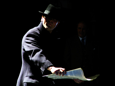 With deep shadows on left, Waist up shot of Philippe, body in profile, holding out a newspaper, head turned out towards audience.