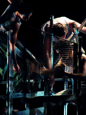 Two separate towers of scaffolding now joined together to create a complex pattern of bars and levels. Three figures are caught in the framework, hanging limply over bars, with blue lighting picking out the steel and the flesh tones of bare arms.