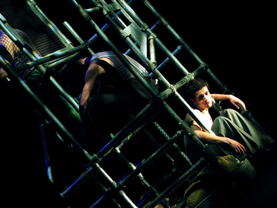 Angle perspective on the scaffold towers in a block centre stage, with young Jacques sitting in the bars and three figures sitting above him in prison vests.