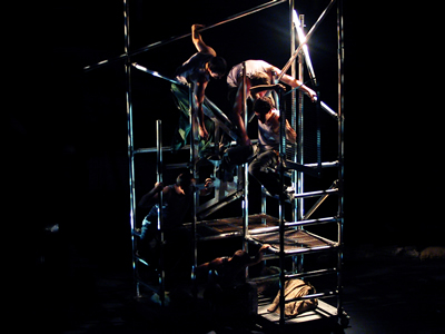 Distant shot of scaffold towers joined to create a block, with five figures hanging within it at different levels, steel and flesh highlighted by blue greeny lighting.