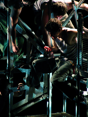 Close up of figures entwined in the scaffold block, against a dark background with a light from above falling on the metal bars and bare flesh.